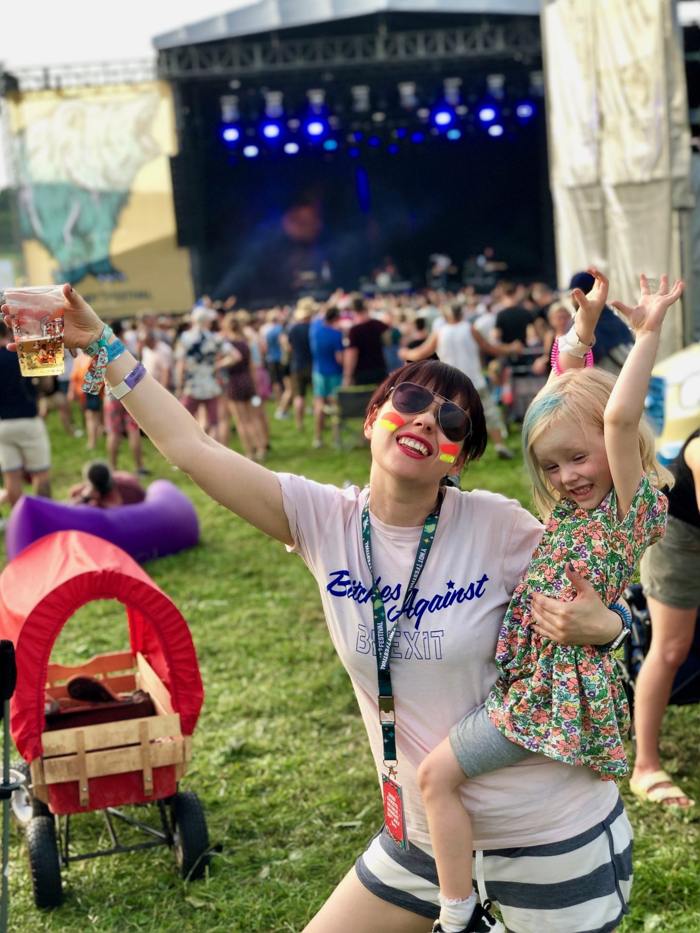Woman celebrates with a pint of lager in one hand and a 4 year old girl on her hip in front of the main stage at a Y Not Festival 2019 - UK biggest festivals.