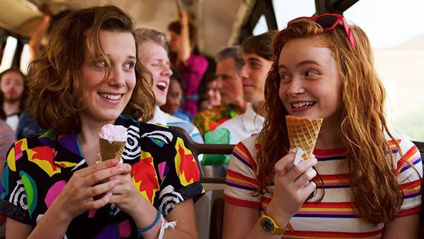 Season 3 Eleven and Max smile whilst eating ice creams on a bus. They are wearing bright clothing. 