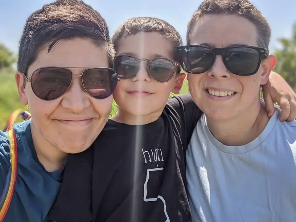 Two women wearing sunglasses hugging a boy. All are smiling. 