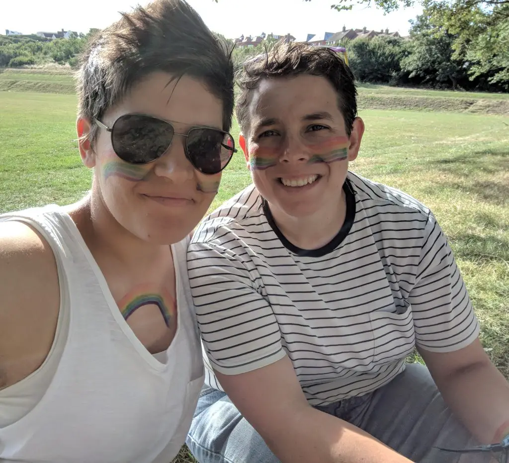 Two woman sat together in a field. Both have rainbow facepaint on and both are smiling.
