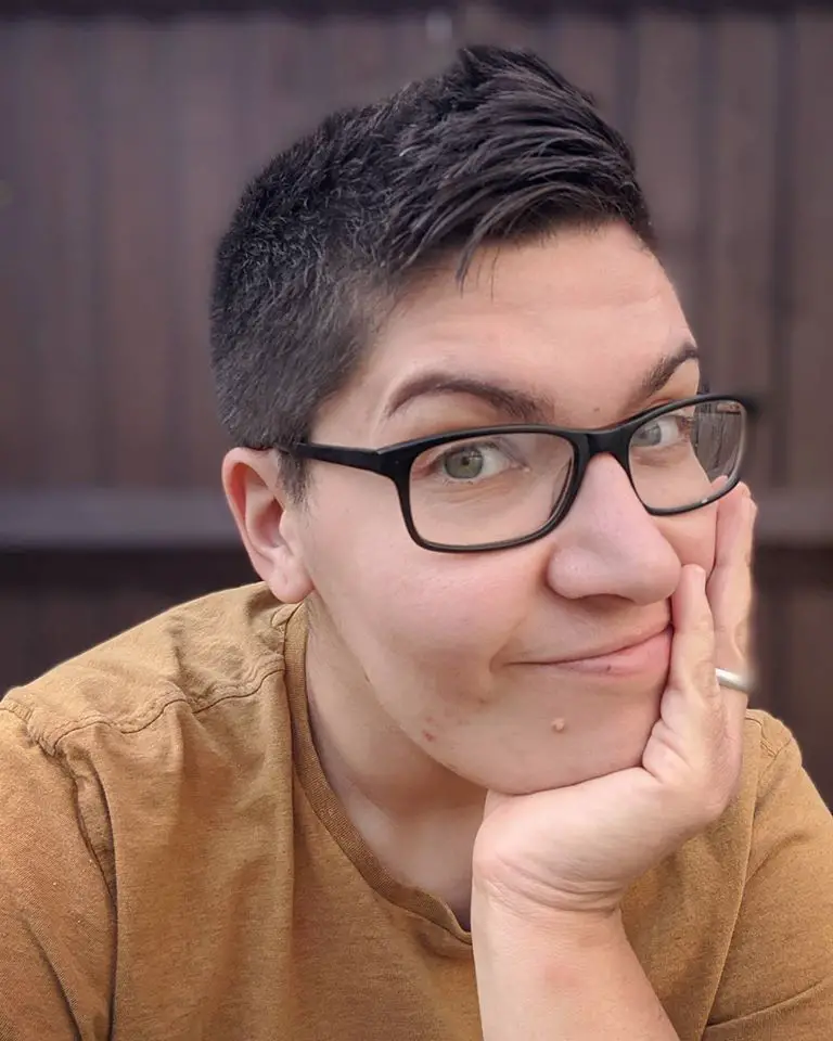 Woman with short brunette hair, wearing glasses and smiling with her hand on her face.