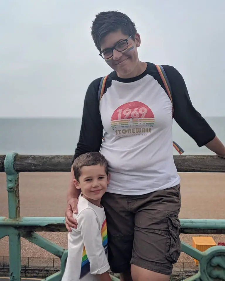 Woman with rainbow facepaint and a 1969 Stonewall t shirt hugging a boy stood at the beach. 