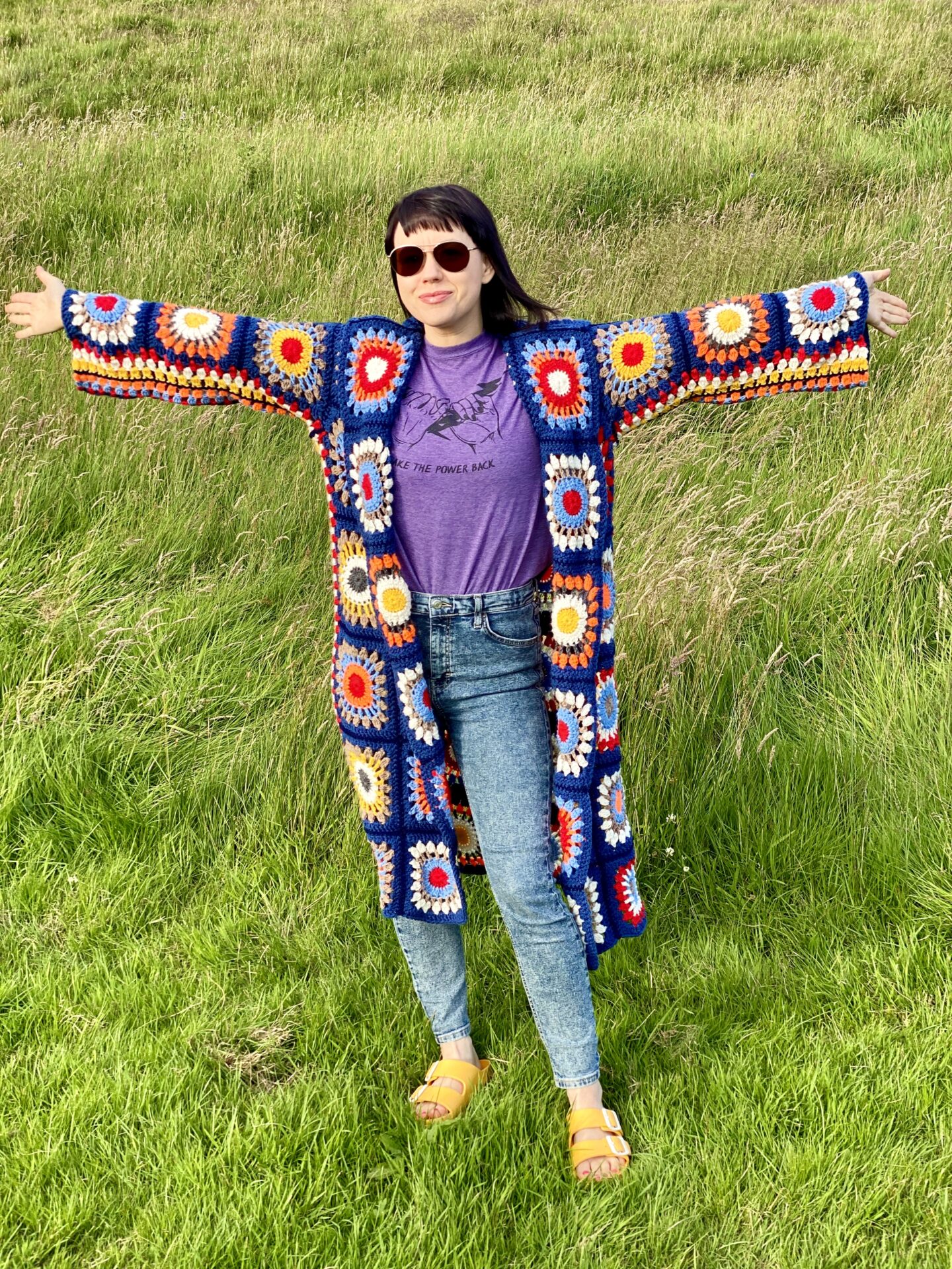 A brunette woman wearing sunglasses and a colourful 70s style crochet Rebecca Cardigan stood in a field with her arms spread wide. 