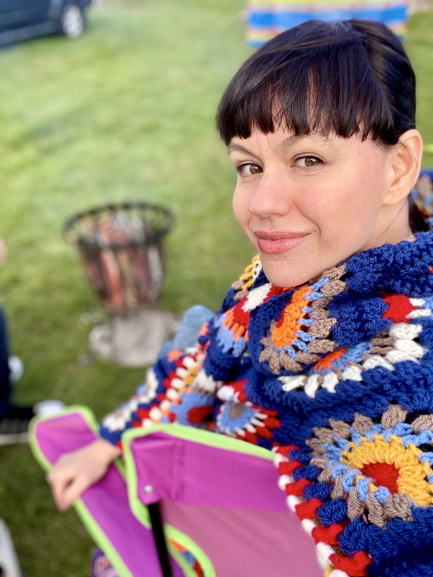 Brunette woman sat in front of a camp fire wearing a colourful crochet Rebecca Cardigan from This Is Us. 