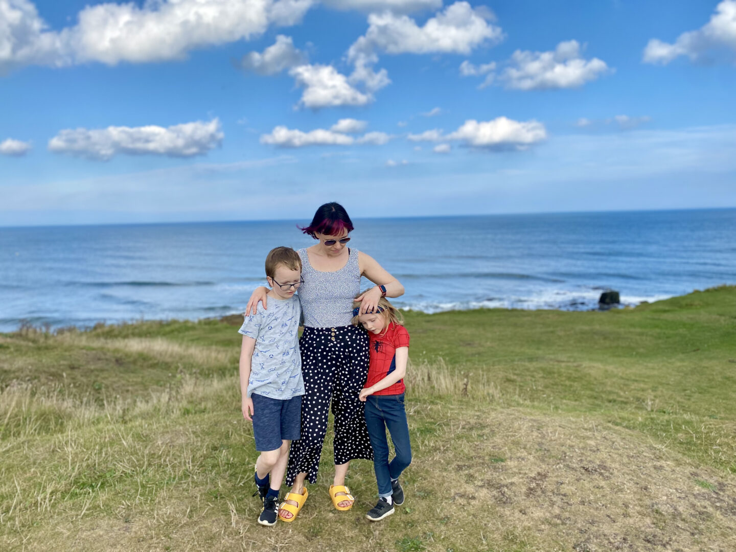 A mam cuddles her son and daughter standing on a cliff top overlooking the sea