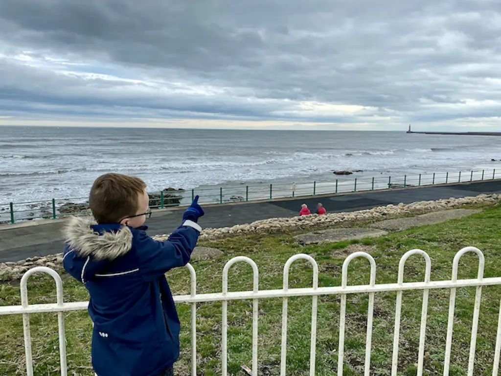 A boy points stands in front on the sea and points at a light house in the distance