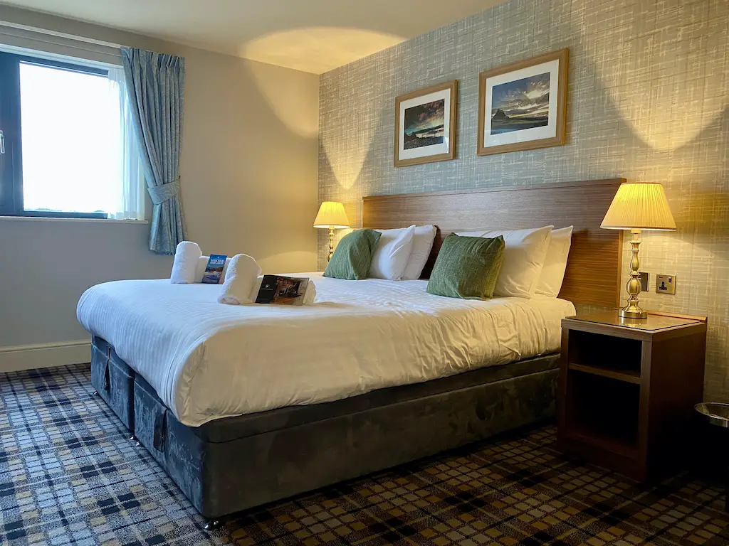 A king size bed with pillows on it and folded towels. Two lamps are glowing on the bedside tables.