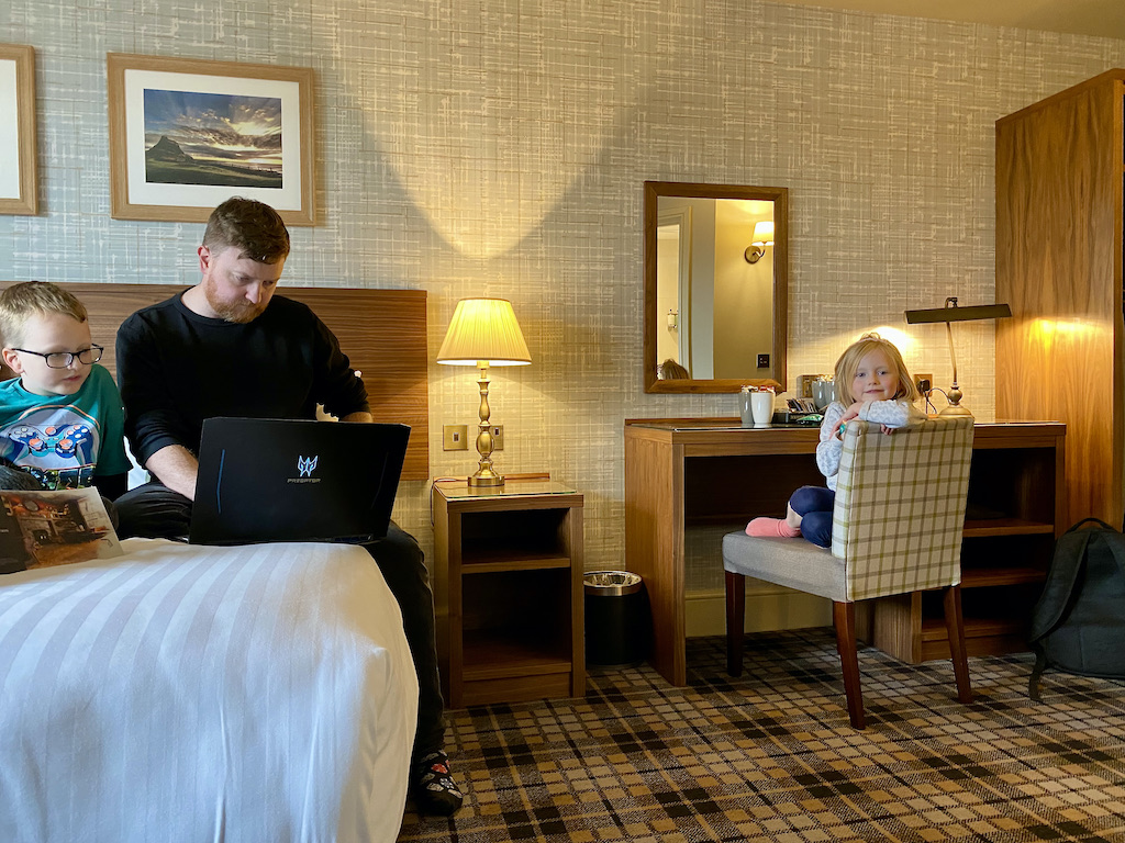 Two boys watch a laptop on a bed and a little girl sits at a desk
