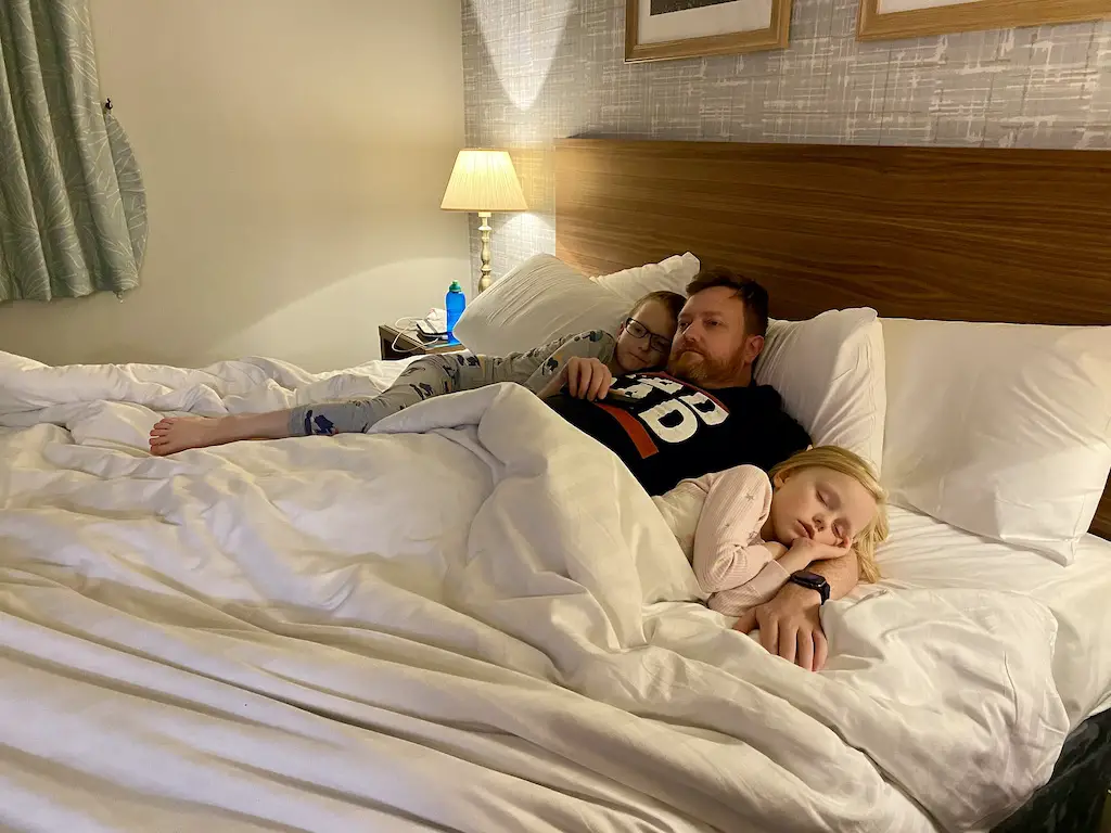 A man cuddles his son and daughter in a king size bed. The girl sleeps on his arm.
