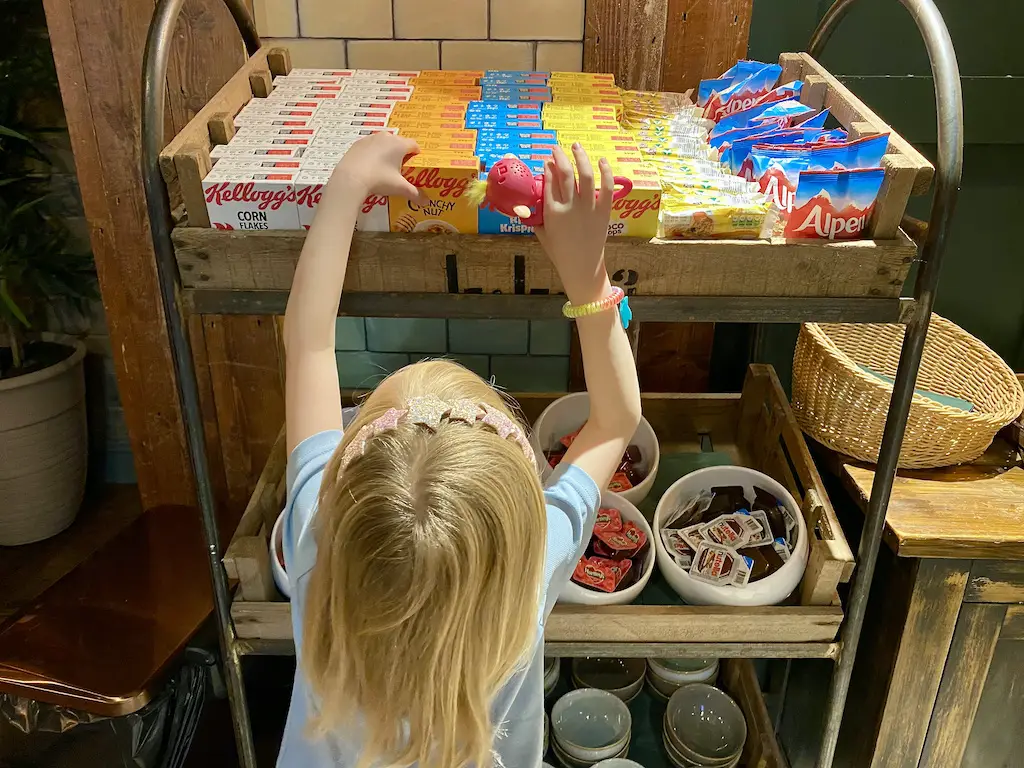 A little girl with blonde hair reaches for breakfast cereal boxes on a breakfast trolley
