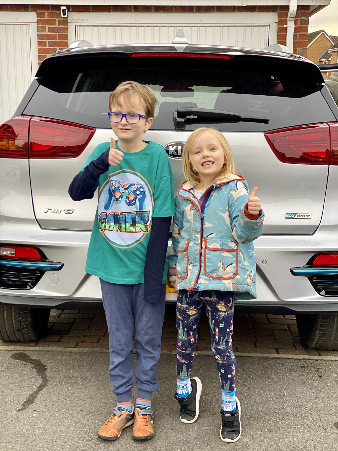 2 children smile with their thumbs up in front of a silver electric car