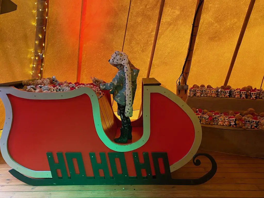 A child stands in a red wooden sleigh with a sign that says ho ho ho as they choose a teddy bear from a large selection. 