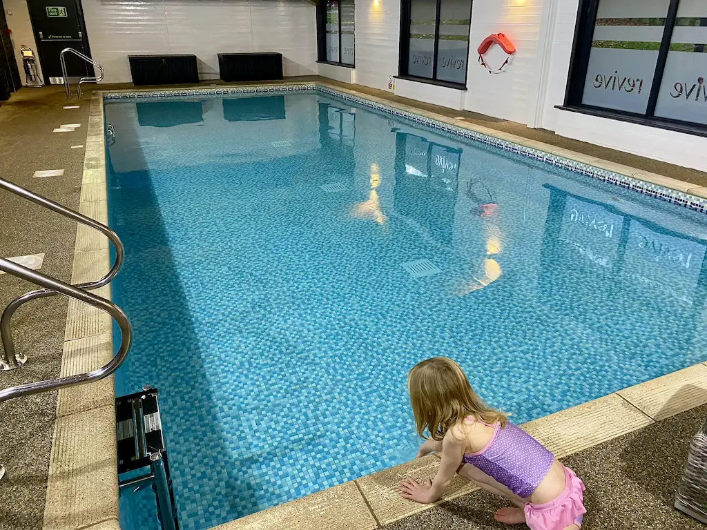 A little blonde girl wearing a purple and pink swim suit touches the water at a swimming pool. 