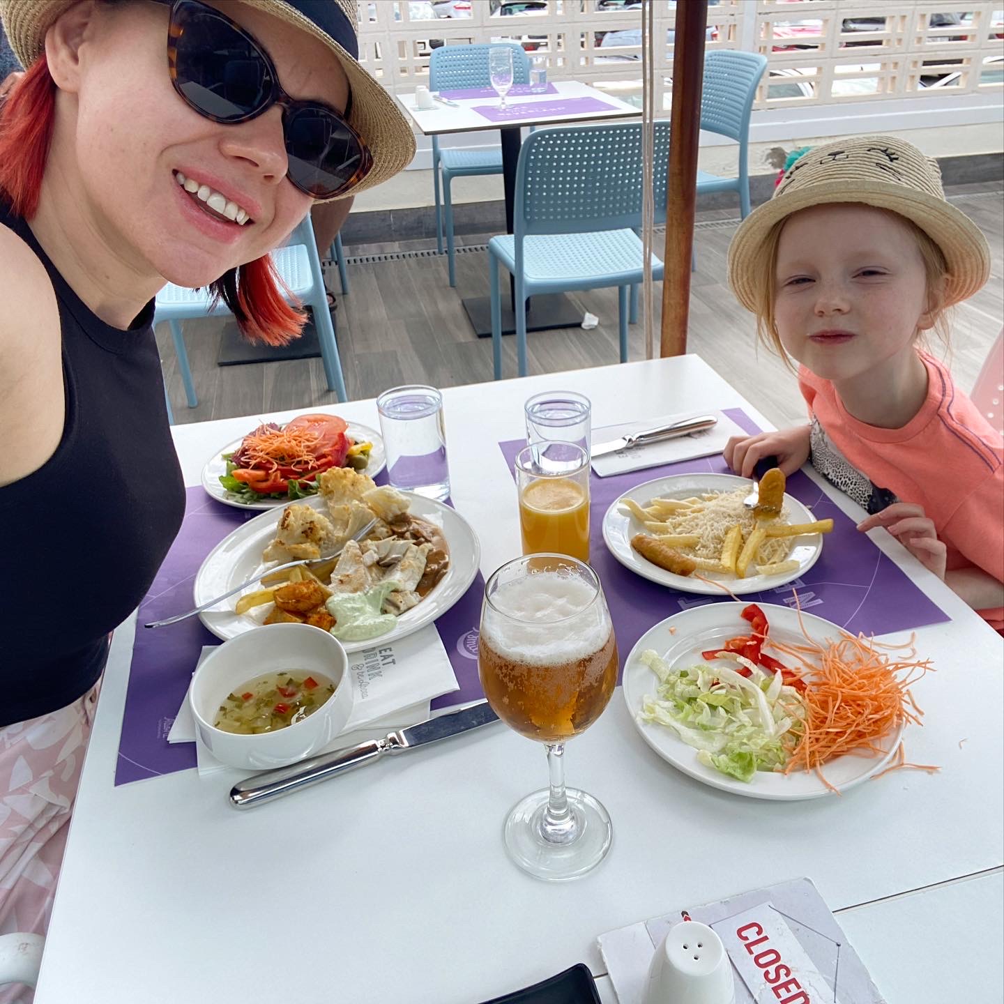 Mother and daughter smile at the camera as they eat lunch at an outdoor restaurant. 