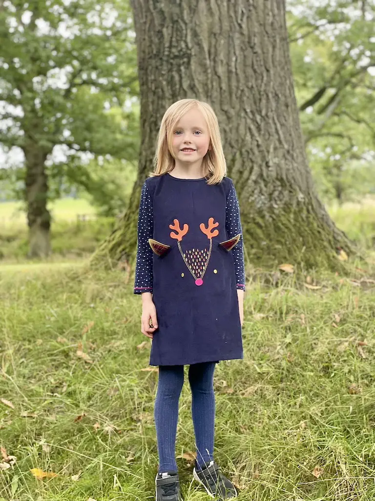 Little girl with blonde hair stood in front of a tree wearing a navu blue courdroy dress with a reindeer on it