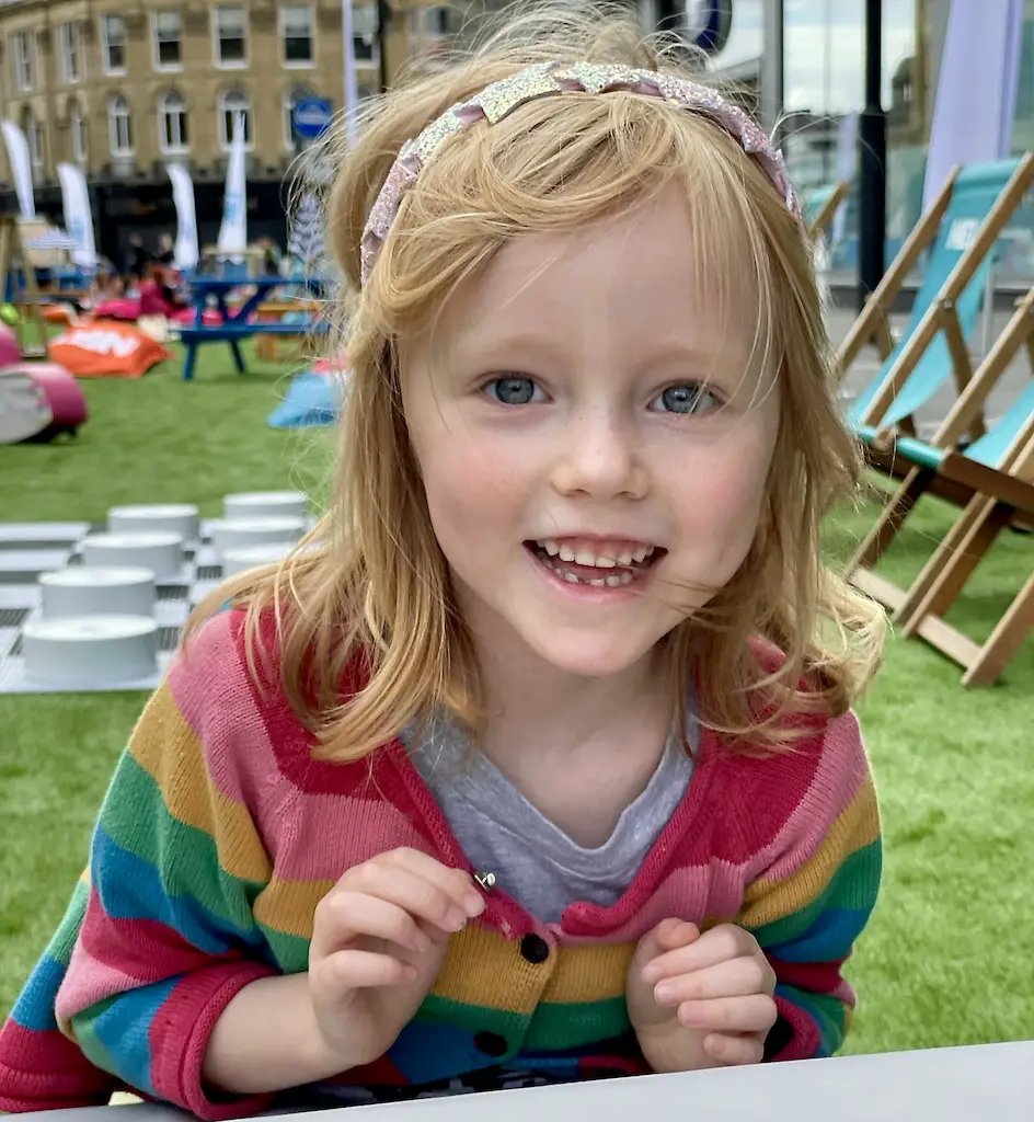 Little girl with blonde hair smiling, wearing a rainbow coloured cardigan