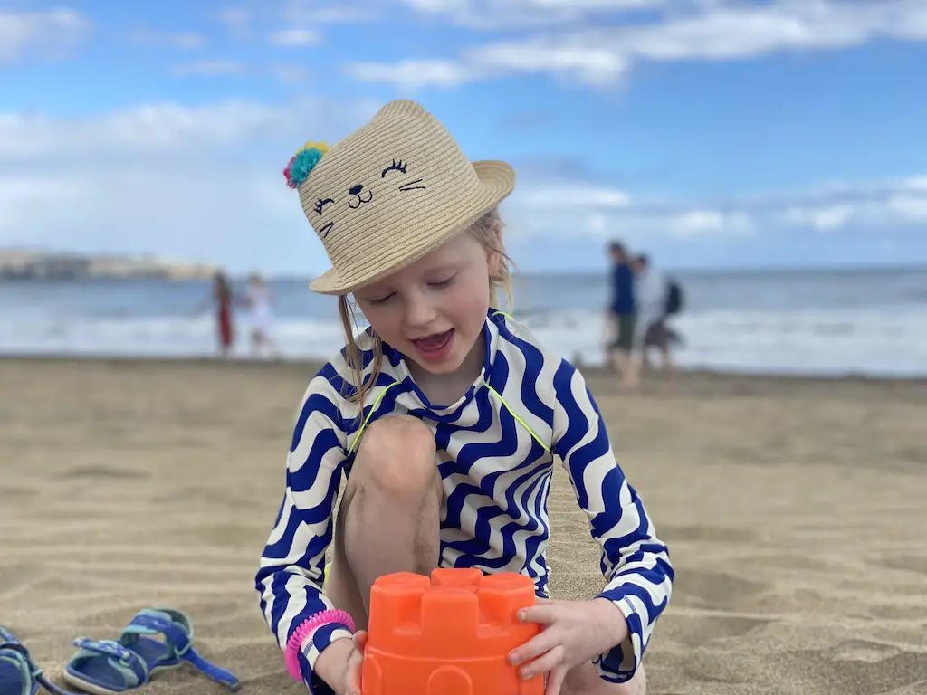 A little girl wearing a sun hat and blue and white stripey swim top plays with a bucket at the beach