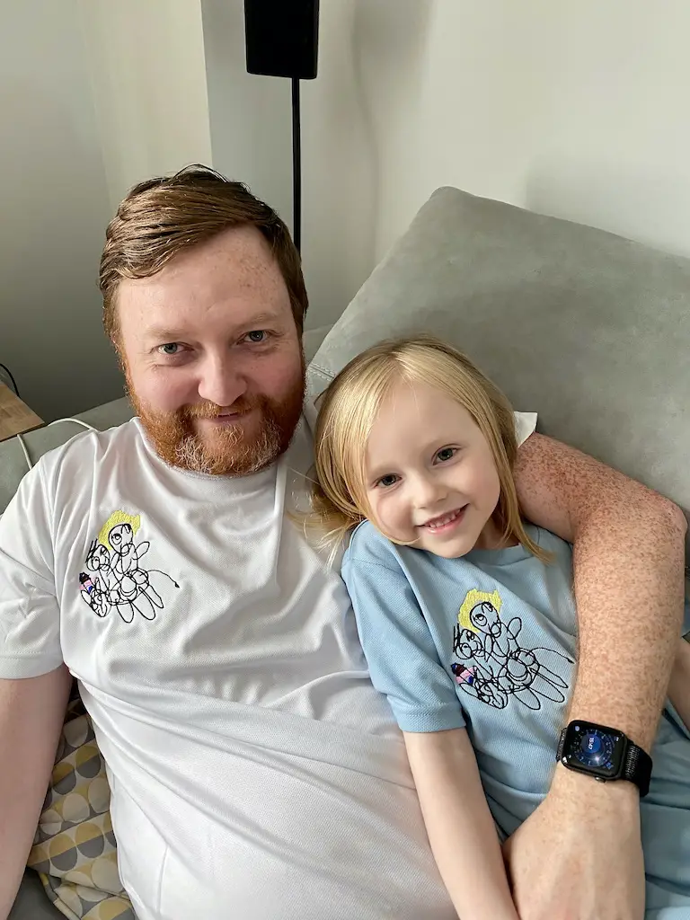 Father and daughter smile at the camera wearing matching t-shirts with a child's drawing on them 