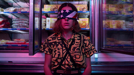 Stranger Things Season 3 Eleven wears a yellow top with black patters and crossed suspenders and an american flag blindfold