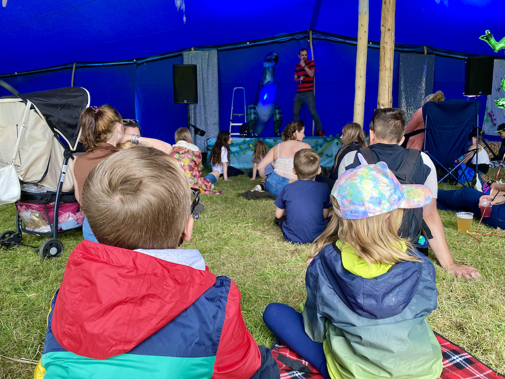 Children watching a man perform a comedy show on a stage in an outdoor tent. 