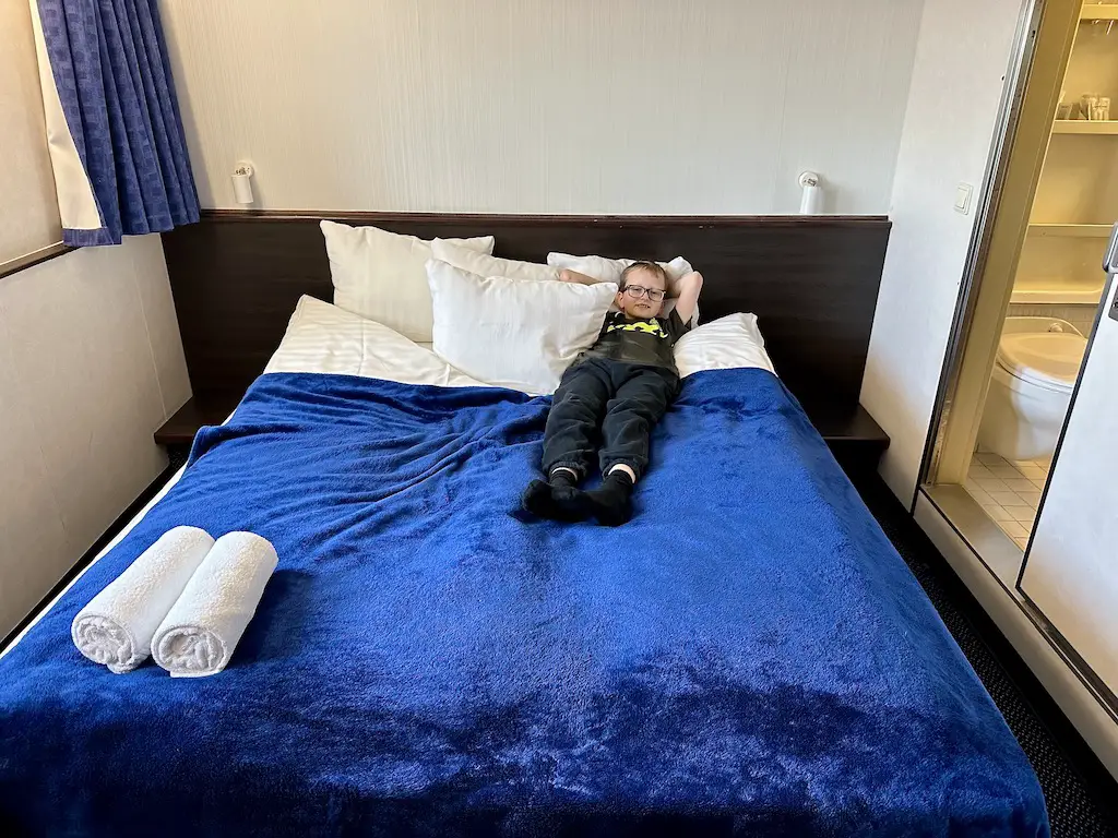 little boy lays in a large double bed with a blue blanket in a Commodore cabin on a DFDS mini cruise