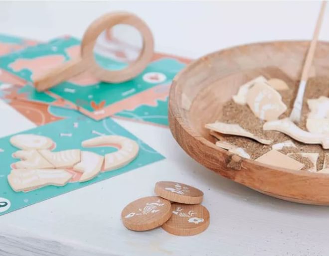 A wooden bowl with sand and small wooden dino pieces, a wooden magnifying glass