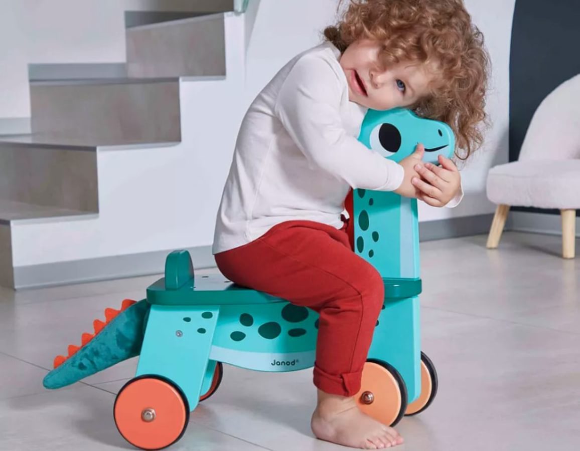 A large turquoise wooden dinosaur ride on toy. A child is sat on it hugging the dino. 