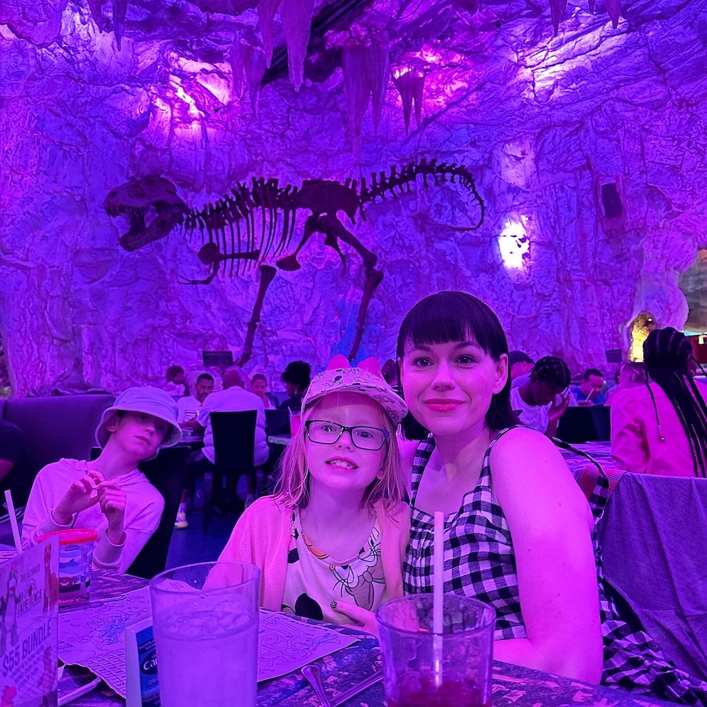A mother and daughter at T Rex restaurant at Disney Springs. There is a T Rex skeleton in the background and the photo has a purple hue. 